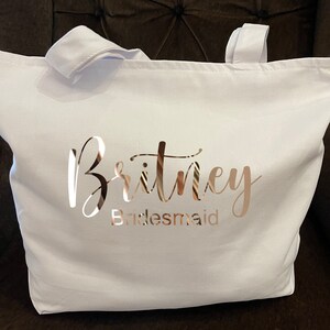 May include: A white tote bag with two white handles. The bag features the name "Britney" in a cursive font and the word "Bridesmaid" in a block font, both in a metallic rose gold color. The bag is made of a sturdy fabric.