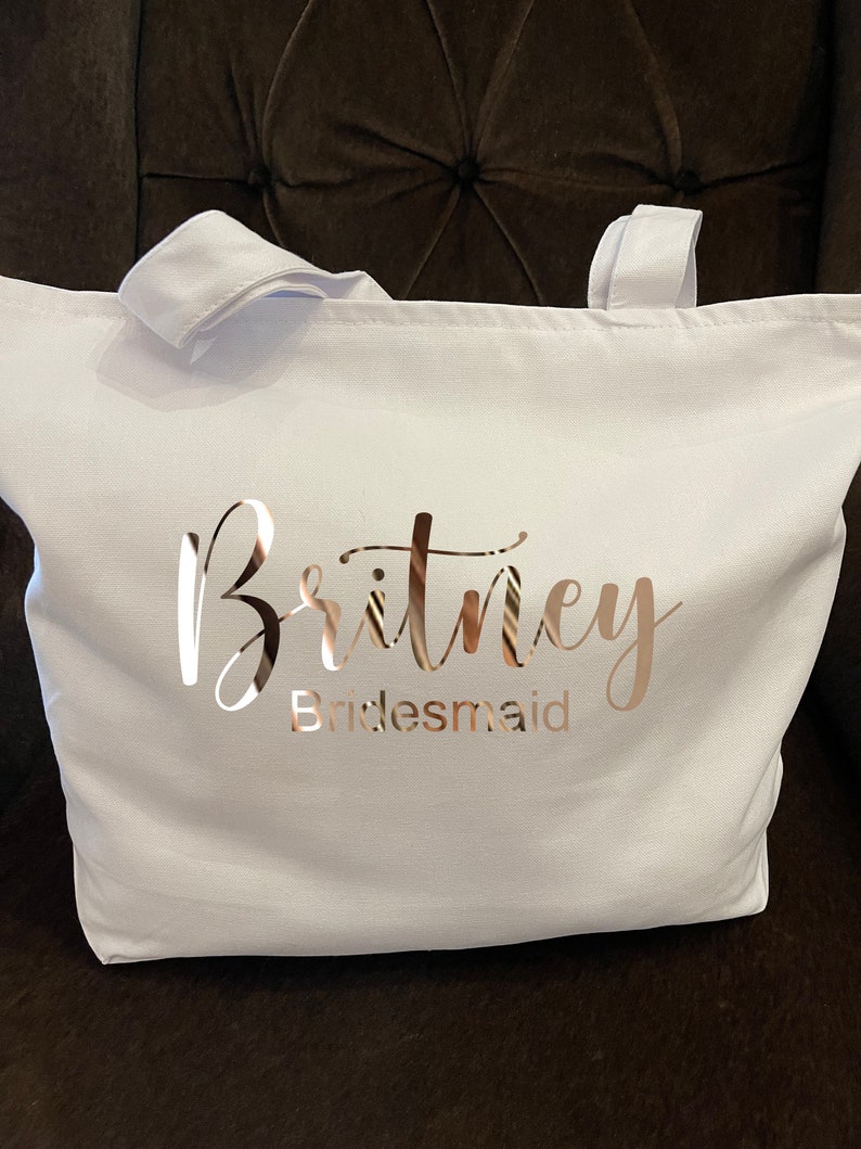 May include: A white tote bag with two white handles. The bag features the name "Britney" in a cursive font and the word "Bridesmaid" in a block font, both in a metallic rose gold color. The bag is made of a sturdy fabric.