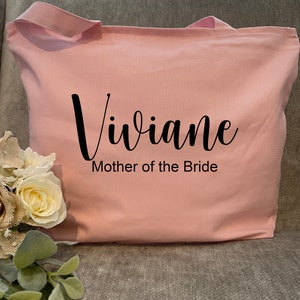 May include: A light pink tote bag with the name "Viviane" in black script and the text "Mother of the Bride" printed below. The bag has two handles and is made of a soft fabric. A bouquet of white and cream-colored flowers is visible on the left.