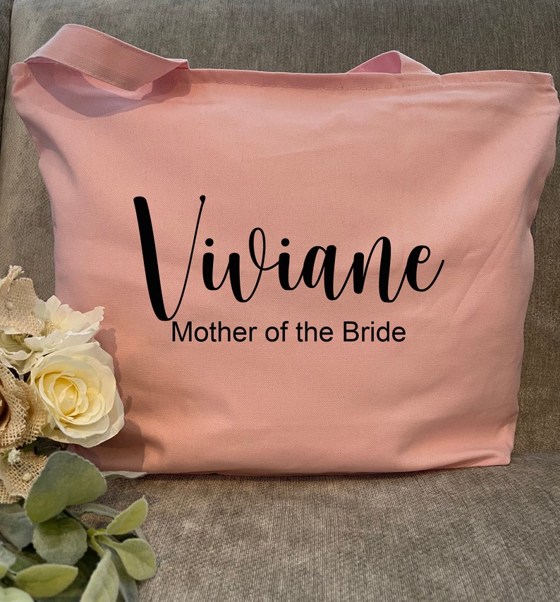 May include: A light pink tote bag with the name "Viviane" in black script and the text "Mother of the Bride" printed below. The bag has two handles and is made of a soft fabric. A bouquet of white and cream-colored flowers is visible on the left.