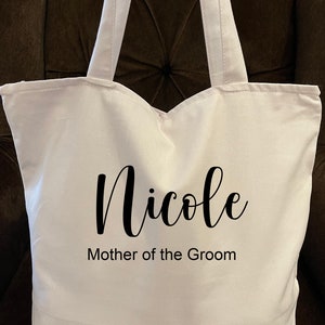 May include: A white tote bag with two handles. The bag has the name "Nicole" in a cursive font, with "Mother of the Groom" printed below it in a simple font. The bag is made of a sturdy fabric.