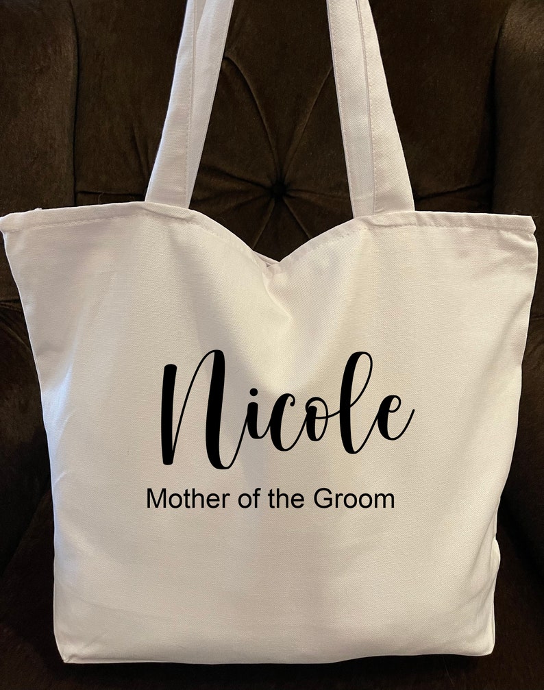 May include: A white tote bag with two handles. The bag has the name "Nicole" in a cursive font, with "Mother of the Groom" printed below it in a simple font. The bag is made of a sturdy fabric.