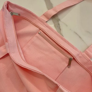 May include: A light pink canvas tote bag with a zippered main compartment and an exterior zippered pocket. The bag has matching pink handles and a silver zipper pull. The bag is made of a sturdy fabric.