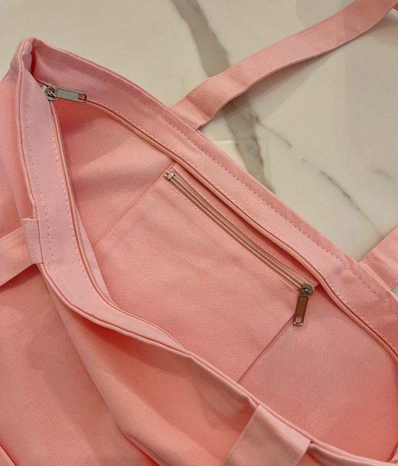 May include: A light pink canvas tote bag with a zippered main compartment and an exterior zippered pocket. The bag has matching pink handles and a silver zipper pull. The bag is made of a sturdy fabric.