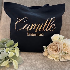May include: Black tote bag with gold lettering that reads "Camille Bridesmaid." The bag is next to a bouquet of white and tan flowers and greenery. The bag has two handles.