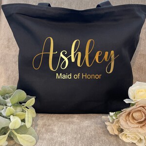 May include: Black tote bag with gold lettering that reads "Ashley Maid of Honor." The bag has two black handles and is made of a sturdy fabric. The bag is decorated with floral accents.