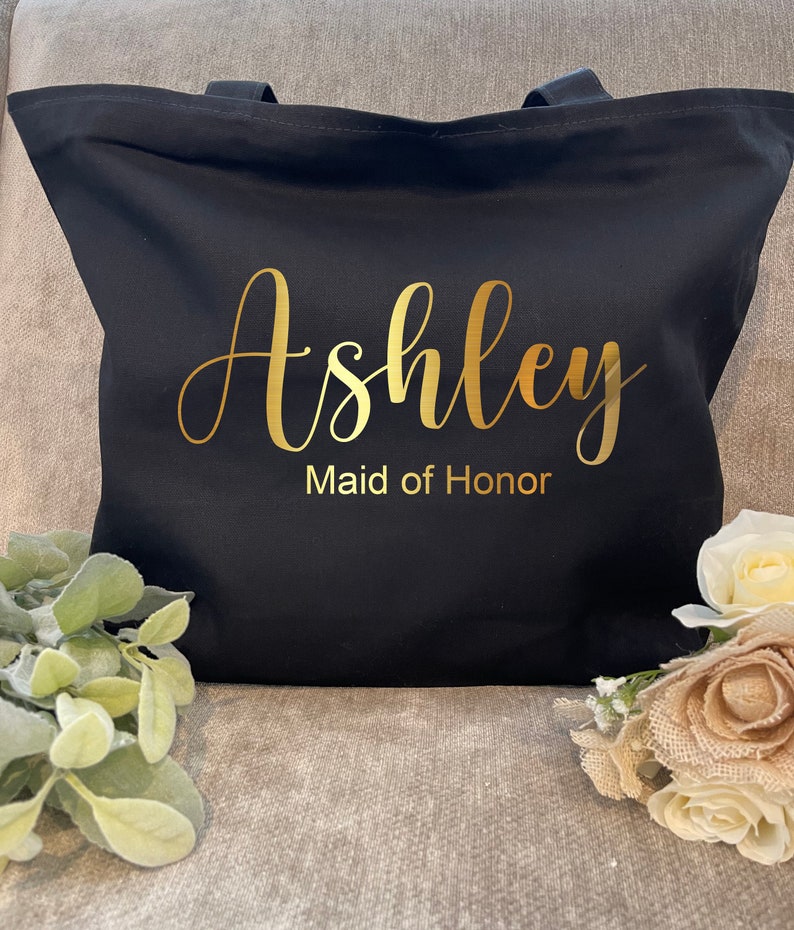 May include: Black tote bag with gold lettering that reads "Ashley Maid of Honor." The bag has two black handles and is made of a sturdy fabric. The bag is decorated with floral accents.