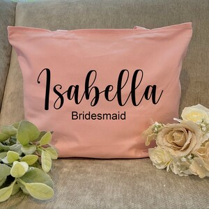 May include: A light pink tote bag with the name "Isabella" in a black script font and the word "Bridesmaid" below it. The bag is sitting on a gray surface next to a bouquet of flowers and greenery.