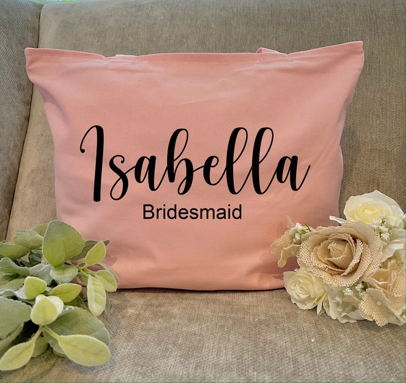 May include: A light pink tote bag with the name "Isabella" in a black script font and the word "Bridesmaid" below it. The bag is sitting on a gray surface next to a bouquet of flowers and greenery.
