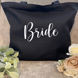 May include: A black tote bag with the word "Bride" in white script. The bag has two handles and is made of a sturdy fabric. The bag is sitting on a gray surface, with greenery and flowers in the foreground.