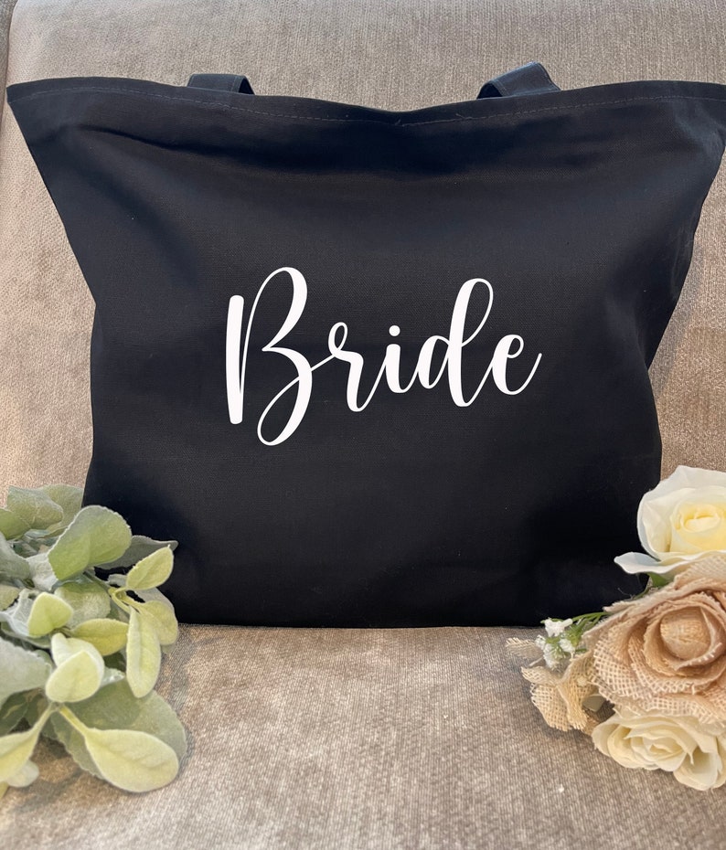 May include: A black tote bag with the word "Bride" in white script. The bag has two handles and is made of a sturdy fabric. The bag is sitting on a gray surface, with greenery and flowers in the foreground.
