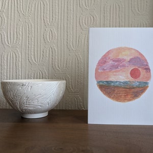 May include: A white ceramic bowl with a fern leaf pattern stands on a wooden surface beside a greeting card. The card shows a watercolour painting of a sunset over the sea, with pink and orange colours.