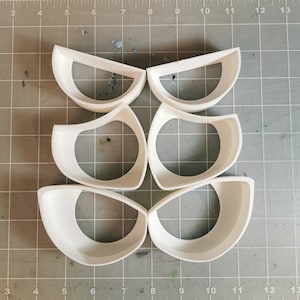 Puede incluir: Seis moldes de plástico blanco en forma de semicírculo. Los moldes están dispuestos en un patrón sobre una superficie de rejilla gris.