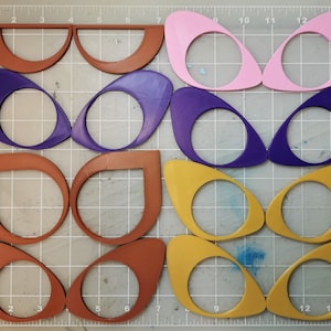 May include: A set of 12 die-cut shapes in various colours, including brown, purple, pink, and gold. The shapes are all similar in design, with a rounded, oval-like form and a hole in the centre. The shapes are arranged in three rows of four, with each row featuring a different colour.