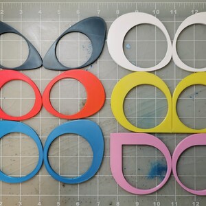 May include: A set of 10 colorful, oval-shaped, cutout shapes with a circular hole in the center. The shapes are arranged in two rows of five, with each row featuring a different color: black, red, blue, white, and yellow.
