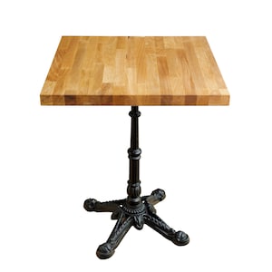 May include: A square wooden table top with a black cast iron base. The table top is made of light wood and has a natural finish. The base has a decorative design with four legs.