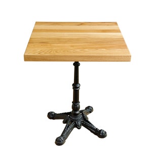 May include: Square wooden table with a light brown tabletop and a black cast iron base. The tabletop is made of wooden planks. The base has a decorative, ornate design. The table is suitable for a cafe or bistro setting.
