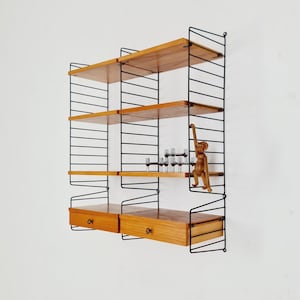 May include: A wall-mounted shelving unit with a black metal frame and wooden shelves and drawers. The shelves are made of light-coloured wood. A small wooden monkey figurine and several small glasses are on the shelves.