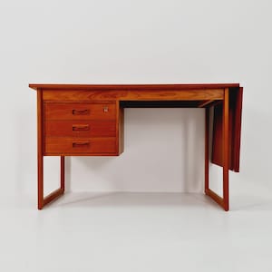 May include: A mid-century modern wooden desk with a warm, reddish-brown finish. The desk features three drawers on the left side, a keyhole, and a drop-leaf extension on the right. The desk has a minimalist design with clean lines and a rectangular shape.