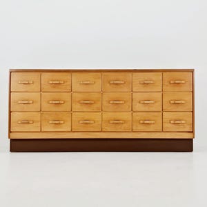 May include: A light-colored wooden cabinet with twenty drawers. The cabinet has a dark brown base and a light-colored wooden frame. Each drawer has a small, curved handle. The cabinet is against a white background.