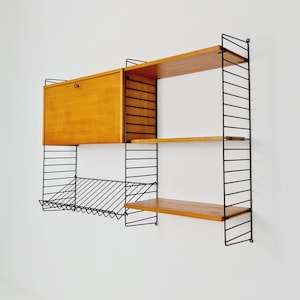May include: A vintage wall-mounted shelving unit with a wooden cabinet and shelves. The light brown wooden elements are supported by a black metal wire frame. The unit includes a magazine rack and is mounted on a white wall.