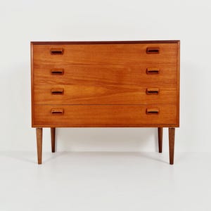 May include: A medium-brown wooden chest of drawers featuring four horizontal drawers, each equipped with two rectangular recessed handles. The furniture piece is supported by four tapered cylindrical legs and displays a visible wood grain pattern.