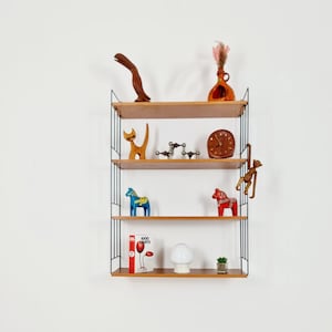 Puede incluir: Una estantería de pared de metal negro con cuatro estantes de madera. Los estantes están decorados con varios artículos, incluyendo un pájaro de madera, un gato de madera, un reloj, un libro, una lámpara y una pequeña planta en maceta.