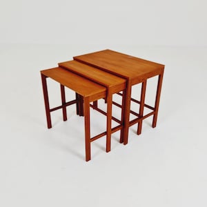 Puede incluir: Un juego de tres mesas nido fabricadas en madera de tono medio. Cada mesa presenta un tablero rectangular y cuatro patas rectas conectadas por barras de soporte laterales, lo que permite que las mesas más pequeñas se deslicen bajo la mayor.