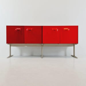 May include: A contemporary red cabinet with four doors and silver handles. The cabinet is raised on silver legs. The doors have a glossy finish, reflecting the light. The cabinet is against a white background.