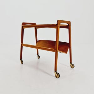 May include: A mid-century modern serving cart made of wood, featuring two tiers and a lower shelf. The cart has a rectangular shape with rounded corners and is supported by four legs with brass-colored casters. The wood has a warm, reddish-brown tone.