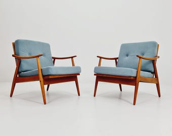May include: Two mid-century modern armchairs. Each chair has a light blue cushion and backrest, with a wooden frame and armrests. The chairs are set against a white background.