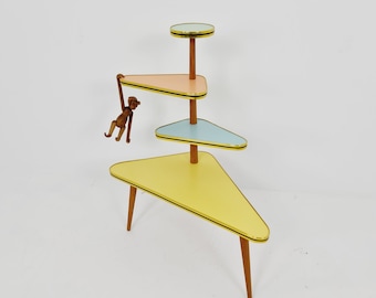 May include: A three-tiered plant stand with a triangular base and shelves. The stand is made of wood and has a yellow, blue, and pink colour scheme. A wooden monkey figurine hangs from the top shelf.