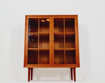 May include: A mid-century modern wooden display cabinet with glass doors. The cabinet has a rectangular shape with a warm brown finish. It features two glass doors with multiple rectangular panes and sits on four tapered legs.