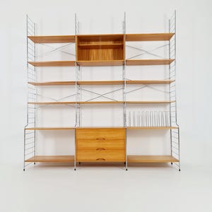 Puede incluir: Un estante modular con estantes y cajones de madera clara, soportado por una estructura metálica. La unidad incluye estantes abiertos, un armario y una sección con divisores verticales. La madera tiene un tono cálido y natural.