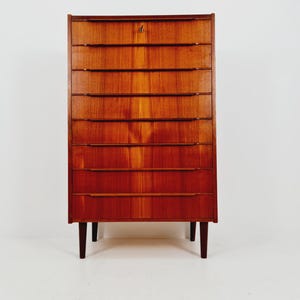 May include: A tall, mid-century modern wooden dresser with eight drawers. The dresser is made of warm-toned wood and has a keyhole at the top. It stands on four slender, dark-colored legs.