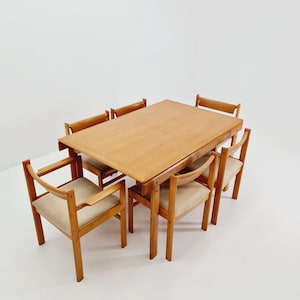 Könnte beinhalten: Ein hellfarbiger Esstisch aus Holz mit sechs passenden Stühlen. Die Stühle haben hell gepolsterte Sitze und geschwungene Armlehnen. Der Tisch und die Stühle stehen vor einem weißen Hintergrund und zeigen ein modernes Design aus der Mitte des Jahrhunderts.