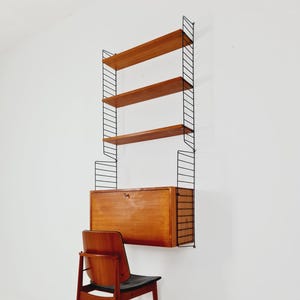 May include: A mid-century modern wall-mounted shelving unit with four wooden shelves and a cabinet. The shelves and cabinet are a warm brown color, supported by a black metal frame. A wooden chair with a black seat is in front of the unit.