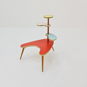 Op de afbeelding: Een mid-century modern plantenstandaard met een rood, roze, blauw en geel kleurenschema. De standaard heeft drie niveaus en een boomerangvormige basis.