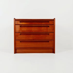 May include: A wooden chest of drawers featuring four horizontal drawers with integrated recessed handles. The piece is constructed from a warm-toned wood with a visible grain pattern and has rounded vertical edges on both the left and right sides.