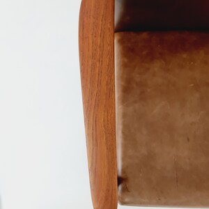 May include: Close-up of a brown leather chair with a wooden armrest. The armrest is curved and has a natural wood grain.