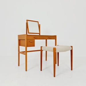 May include: A light brown wooden vanity table featuring a rectangular mirror, two small drawers, and a single side drawer with integrated handles. The vanity is accompanied by a matching wooden stool with a light beige upholstered seat cushion.