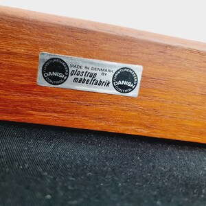 May include: Close-up of a wooden chair with a black fabric seat. The chair has a label that reads "MADE IN DENMARK BY GLOSTRUP MABELFABRIK" and two circular labels that read "FURNITURE MAKERS DANISH CONTROL".