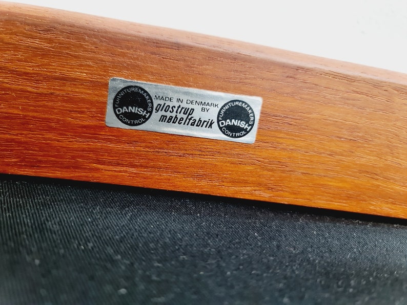 May include: Close-up of a wooden chair with a black fabric seat. The chair has a label that reads "MADE IN DENMARK BY GLOSTRUP MABELFABRIK" and two circular labels that read "FURNITURE MAKERS DANISH CONTROL".