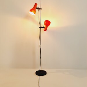 Puede incluir: Una l&aacute;mpara de pie de cromo con dos pantallas ajustables de color naranja. La l&aacute;mpara tiene una base negra y un cable negro.