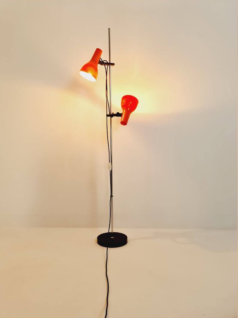 Puede incluir: Una l&aacute;mpara de pie de cromo con dos pantallas ajustables de color naranja. La l&aacute;mpara tiene una base negra y un cable negro.