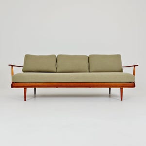 May include: A three-seat sofa featuring a warm-toned wooden frame with tapered legs and curved armrests. The piece includes three rectangular back cushions and a single long seat cushion, all upholstered in a solid light olive green fabric.