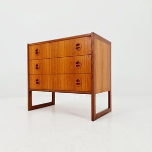 Op de afbeelding: Een houten dressoir in mid-century modern stijl met drie lades. Het dressoir is gemaakt van lichtbruin hout en heeft een rechthoekige vorm. Elke lade heeft twee ronde handgrepen. Het staat op vier rechthoekige poten.