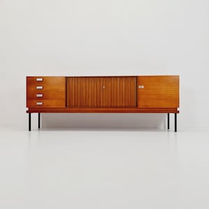 May include: A mid-century modern sideboard with a wood veneer finish and black metal legs. The sideboard has four drawers on the left side and two doors on the right side. The doors have a slatted design.