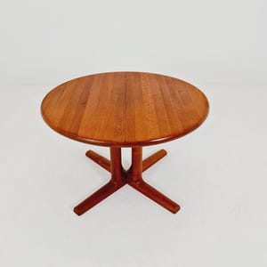 Op de afbeelding: Een ronde houten tafel met een warme, middelbruine houtnerf. Het tafelblad is cirkelvormig en heeft een naad in het midden. De tafel wordt ondersteund door een centrale kolomvoet die uitloopt in vier afzonderlijke, gespreide poten.