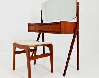 Könnte beinhalten: Ein modernes Schminktisch-Set aus der Mitte des Jahrhunderts mit einem rechteckigen Spiegel, drei Schubladen und einem passenden Hocker. Der Schminktisch und der Hocker sind aus Holz gefertigt und haben einen hellfarbigen Stoffsitz. Der Spiegel ist mit Holzstützen am Schminktisch befestigt.